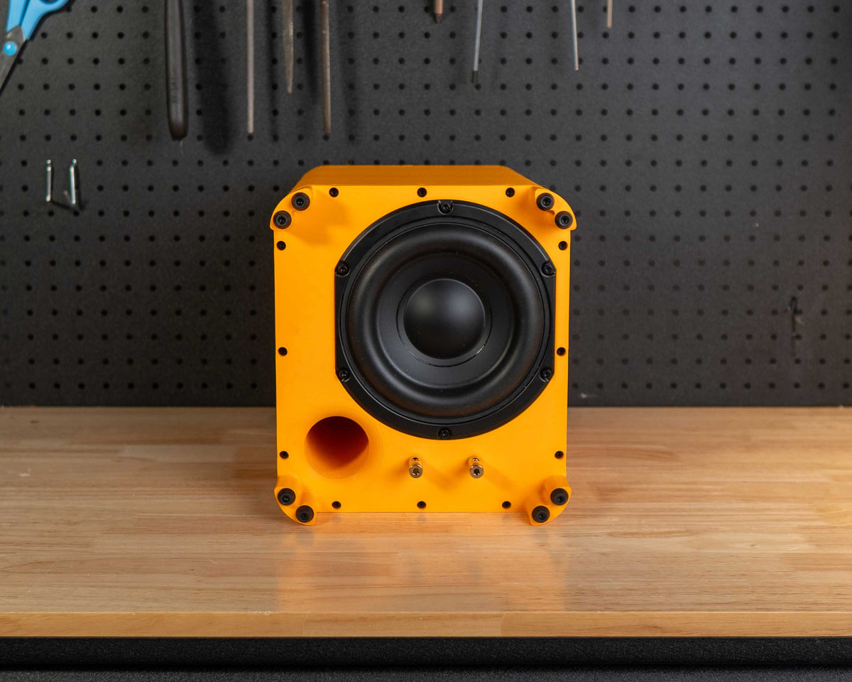 A bright yellow and black speaker subwoofer stands on a wooden workbench in front of a black pegboard workshop wall filled with tools.