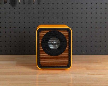 A small square speaker with a yellow-orange frame and black grille sits centered on a wooden workbench in a workshop setting with a black pegboard and tools behind it.