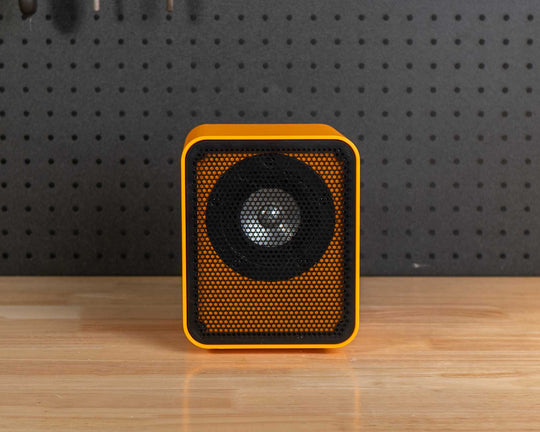 A small square speaker with a yellow-orange frame and black grille sits centered on a wooden workbench in a workshop setting with a black pegboard and tools behind it.