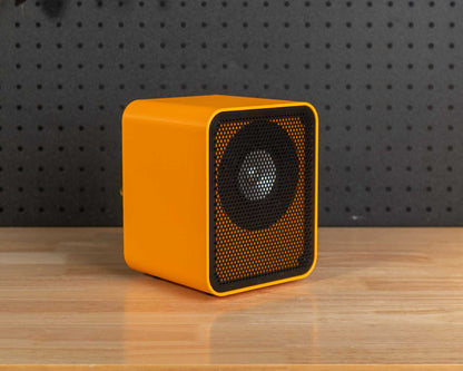 A bright yellow square speaker with a black front grille sits on a wooden workbench in a workshop setting with a black pegboard background filled with tools.