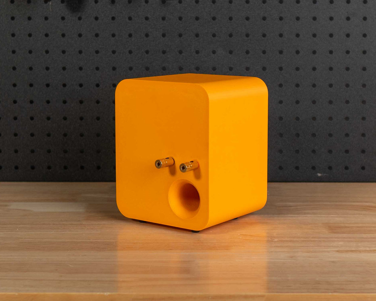 A bright orange cube-shaped speaker with a circular port on the back sits on a wooden workbench in a workshop setting with a black pegboard background.