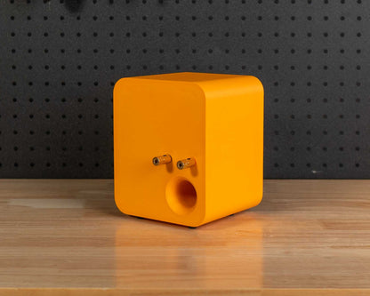 A bright orange cube-shaped speaker with a circular port on the back sits on a wooden workbench in a workshop setting with a black pegboard background.