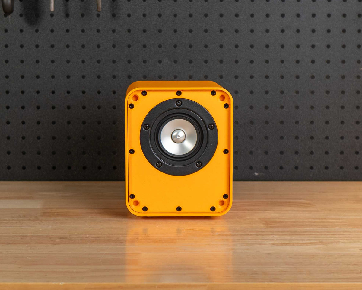 A yellow speaker with a black circular driver sits centered on a light wood workbench in a workshop setting with a black pegboard background filled with tools.